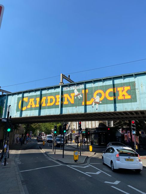 A large blue metal railway bridge with the words 'Camden Lock' painted in bold yellow and red letters spans above a busy pedestrian crossing in Camden town, London. Underneath the bridge, there are nu