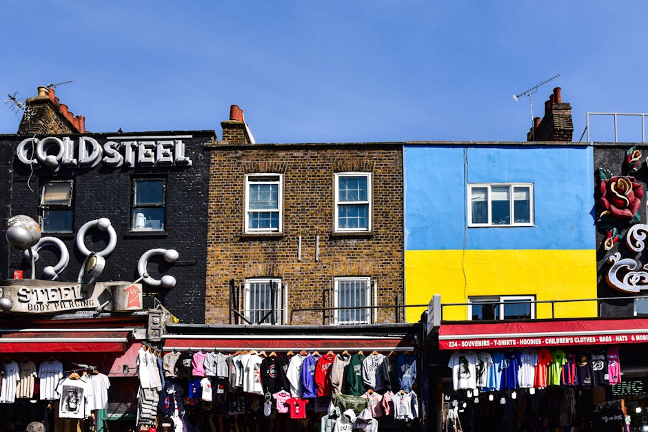 A row of three adjacent buildings on Camden High Street, with the storefront at street level displaying clothing and accessories hanging on racks and hooks, including T-shirts, hoodies, and children's clothes. Above the stores, the first building has a black exterior with graffiti-style art and a sign reading 'COLD STEEL', while the second building has a brown brick façade with three white-framed windows. The third building features a painted exterior with a design resembling the Ukrainian flag, with a blue upper half and a yellow lower half, and a window on the blue section. The street scene is illuminated by daylight, with various objects such as shopping bags, boxes, and packing materials possibly related to home relocation visible on the storefront, and a black metal railing separating the building façade from the sidewalk. This scene illustrates typical urban moving logistics, including the preparation and transportation of household items, as would be relevant in a house removals context on Camden High Street, with Camden Man and Van providing professional removals services.
