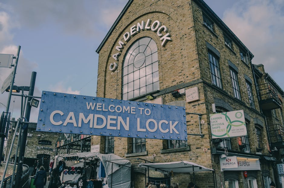 The image depicts a three-story brick building on Camden High Street with large, arched windows and black window frames. Prominently displayed on the building is a blue sign reading 'Welcome to Camden Lock' and another sign with the words 'Camden Lock, Demlock, Shops & Rent,' indicating the location is a commercial area in Camden. In front of the building, several cardboard boxes and plastic-wrapped items are placed on the pavement, suggesting packing materials used during a home relocation or furniture transport. There is also a white tent or canopy covering some stored items, and a metal trolley wheel can be seen, used for moving or lifting heavy objects. The scene is set during daylight with a partly cloudy sky overhead, and the surroundings include street signage, a lamppost, and pedestrians, reinforcing the busy atmosphere typical of a transportation or moving process in an urban area. Camden Man and Van's professional removals services are implied through the context of loading and packing in a tight access location, suitable for typical house or apartment moves on Camden High Street.