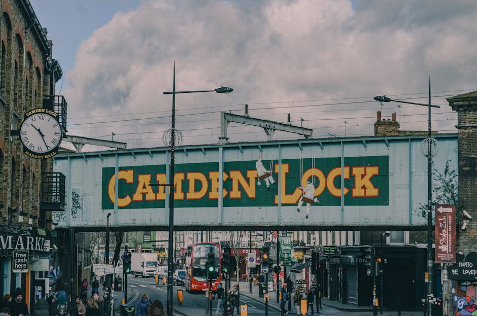 A row of three adjacent buildings on Camden High Street, with the storefront at street level displaying clothing and accessories hanging on racks and hooks, including T-shirts, hoodies, and children's clothes. Above the stores, the first building has a black exterior with graffiti-style art and a sign reading 'COLD STEEL', while the second building has a brown brick façade with three white-framed windows. The third building features a painted exterior with a design resembling the Ukrainian flag, with a blue upper half and a yellow lower half, and a window on the blue section. The street scene is illuminated by daylight, with various objects such as shopping bags, boxes, and packing materials possibly related to home relocation visible on the storefront, and a black metal railing separating the building façade from the sidewalk. This scene illustrates typical urban moving logistics, including the preparation and transportation of household items, as would be relevant in a house removals context on Camden High Street, with Camden Man and Van providing professional removals services.