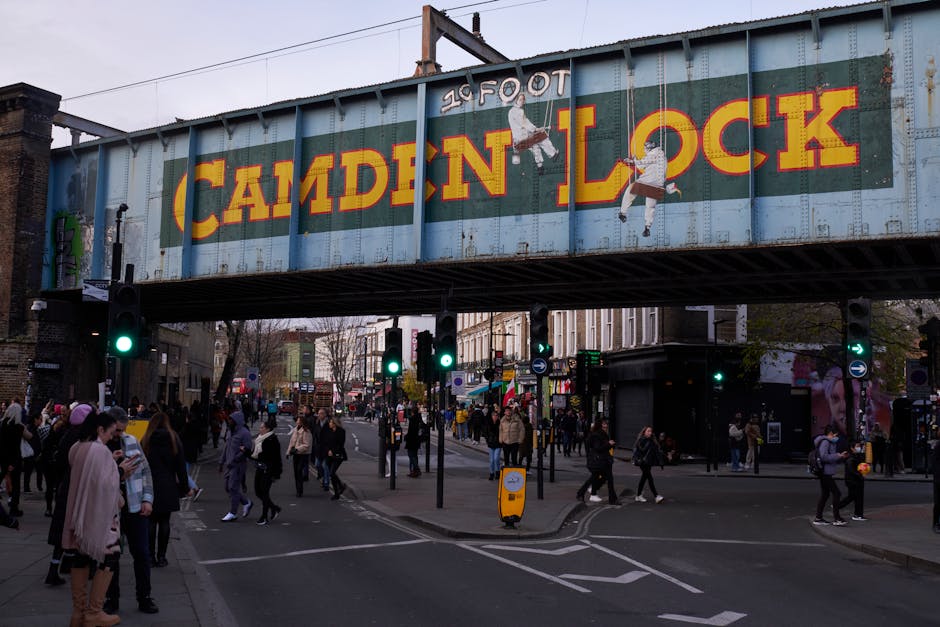 A busy urban street scene beneath a large metal railway bridge with a blue background and bold yellow and red lettering that reads 'Camden Lock.' Pedestrians are crossing the street, some walking and others waiting at traffic lights, on a pavement lined with shops and market stalls. In the foreground, several cardboard boxes and wrapped furniture items are visible, likely part of a home relocation or furniture transport process. Nearby, a large van parked on the side of the road is partly visible, indicating a loading or unloading operation typical of house removals. The lighting suggests late afternoon, with natural light casting shadows on the scene. Camden Man and Van is known for providing removals and moving services, including packing, loading, and transportation, supported by the logistics seen in this lively city environment.