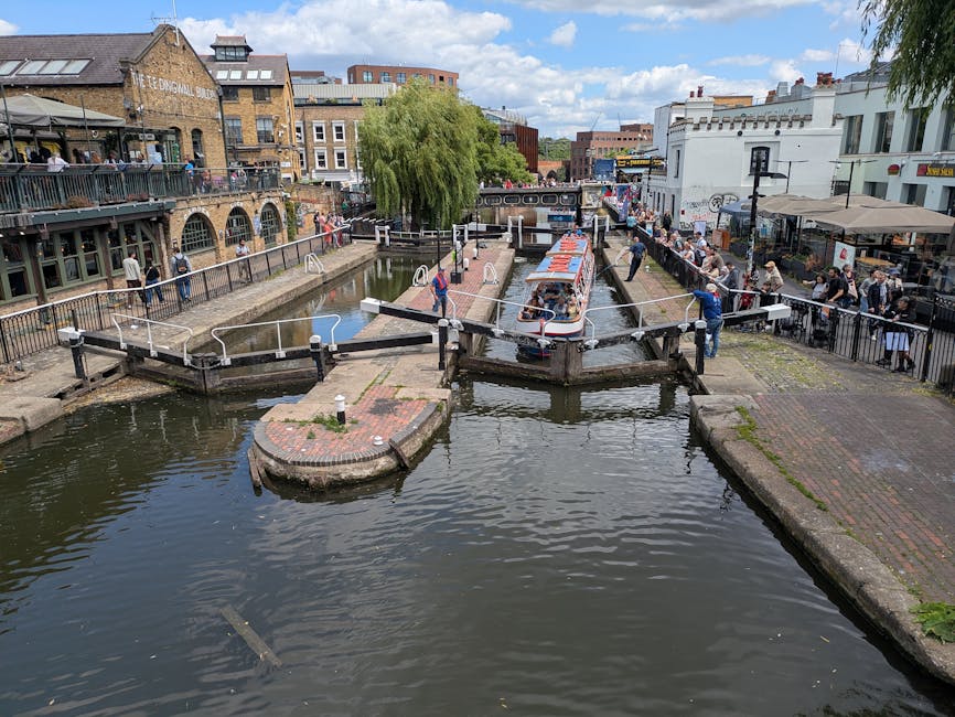 A canal scene in Camden with a passenger boat passing through a narrow lock, surrounded by historic brick and white buildings with outdoor cafes and shops. Several people are walking along the pathways beside the water, some observing the boat and lock operation. The lock gates are open, with metal railings along the edges for safety, and the water level is slightly raised to allow the boat to pass through. The canal is lined with cobblestone and brick pavements. A large green weeping willow tree is visible in the background, and the sky is partly cloudy with patches of blue. This environment illustrates typical urban waterways where house removals and furniture transport could involve logistical planning, loading, and unloading at such locations, while Camden Man and Van's removals services might coordinate with these surroundings during a home relocation or packing process.