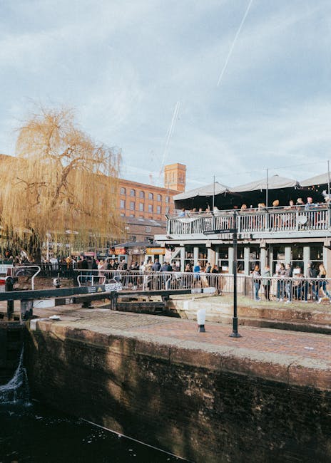 A busy urban street scene beneath a large metal railway bridge with a blue background and bold yellow and red lettering that reads 'Camden Lock.' Pedestrians are crossing the street, some walking and others waiting at traffic lights, on a pavement lined with shops and market stalls. In the foreground, several cardboard boxes and wrapped furniture items are visible, likely part of a home relocation or furniture transport process. Nearby, a large van parked on the side of the road is partly visible, indicating a loading or unloading operation typical of house removals. The lighting suggests late afternoon, with natural light casting shadows on the scene. Camden Man and Van is known for providing removals and moving services, including packing, loading, and transportation, supported by the logistics seen in this lively city environment.