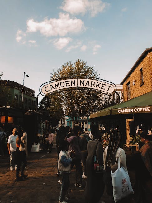 A busy street scene at Camden Market, with numerous pedestrians walking along the pavement and entering shops, including a café with a green awning labeled 'Camden Coffee.' In the background, there is a large arched sign reading 'Camden Market' hanging above the entrance, with trees and a partly cloudy sky overhead. The scene captures the lively atmosphere typical of home relocation and furniture transport activities, as people move through an urban area popular for shopping and socialising. The image emphasizes the environment where house removals and packing initiatives might take place, illustrating the logistical context for moving services offered by companies like Camden Man and Van, including loading and transportation of household items amidst a vibrant market setting.
