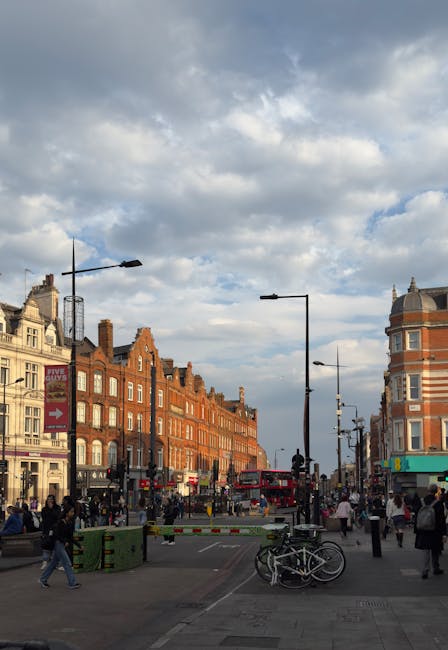 A busy urban street scene in Camden, featuring historic red-brick terraced buildings with multiple windows along the left side, and a prominent rounded corner building with a distinctive dome on the right. The street is filled with pedestrians walking on the pavement, some carrying bags, while others are waiting at the roadway. In the foreground, a set of bicycles is parked near a green bin, and a white and red barrier is positioned across part of the street, possibly indicating a temporary closure or loading zone. Several black street lamps line the street, and overhead, the sky is partly cloudy with patches of blue. The environment suggests a vibrant area with shops, cafes, and possibly a transportation hub nearby. This scene is relevant to house removals and relocating services, highlighting the typical urban setting where furniture transport and home relocation activities occur, as managed by companies like Camden Man and Van during loading or unloading processes.
