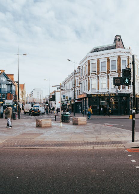 A busy street scene at Camden Market, with numerous pedestrians walking along the pavement and entering shops, including a café with a green awning labeled 'Camden Coffee.' In the background, there is a large arched sign reading 'Camden Market' hanging above the entrance, with trees and a partly cloudy sky overhead. The scene captures the lively atmosphere typical of home relocation and furniture transport activities, as people move through an urban area popular for shopping and socialising. The image emphasizes the environment where house removals and packing initiatives might take place, illustrating the logistical context for moving services offered by companies like Camden Man and Van, including loading and transportation of household items amidst a vibrant market setting.